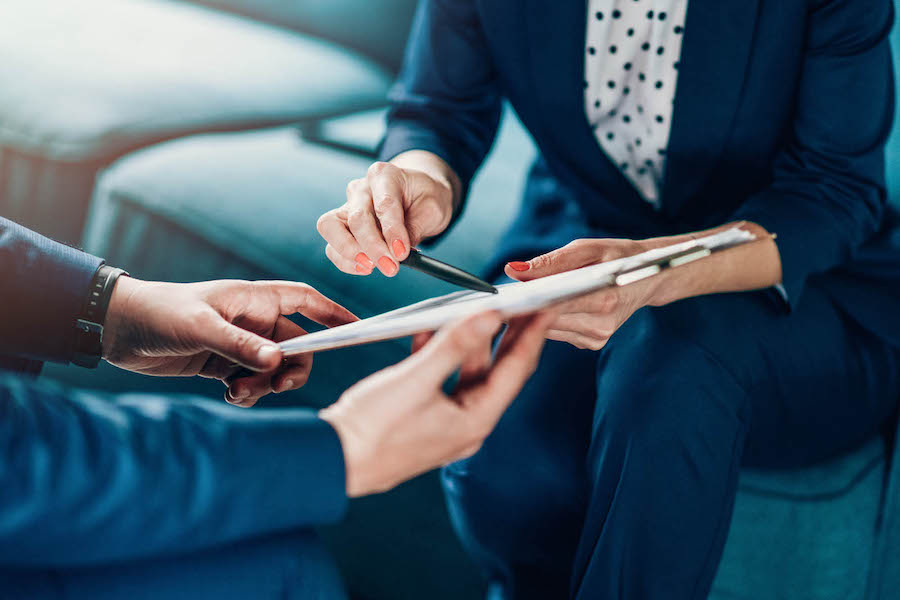 Two business people sit on a couch; one writes notes on paper while the other observes.